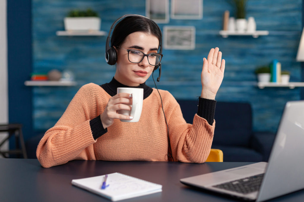 Woman waving during online degree program, studying with laptop and headphones
