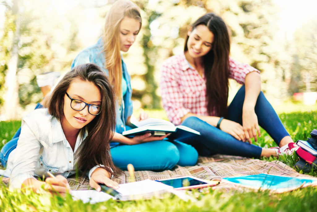Students studying outdoors on grass with books and notebooks.