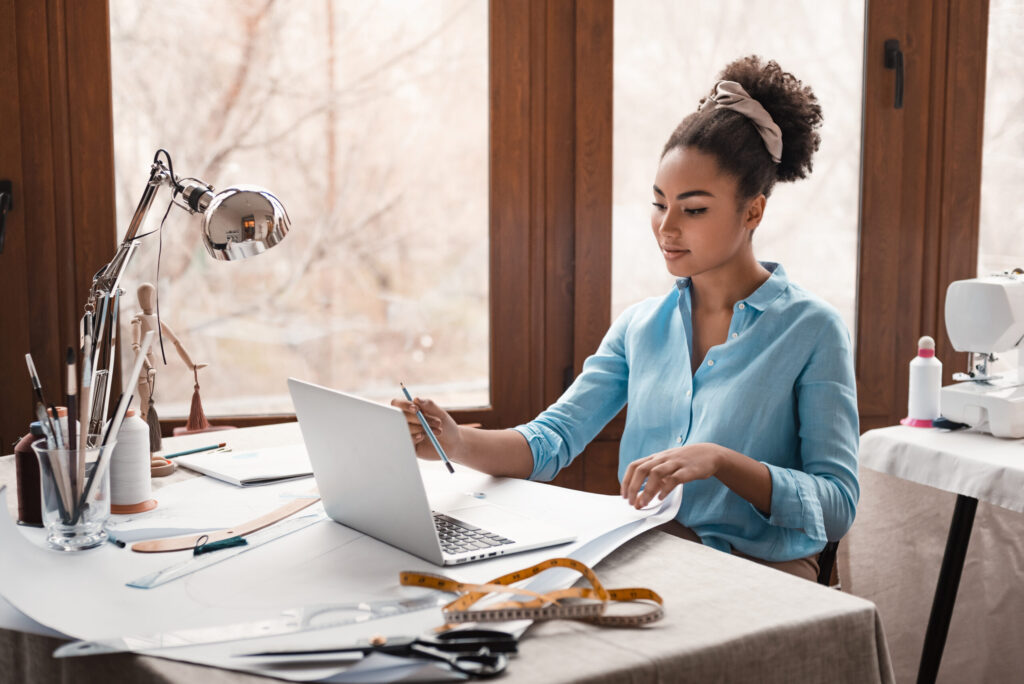 Stylist working on laptop with design plans and sewing supplies for business management.